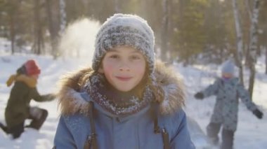 Tatillerde kış ormanlarında poz veren, arka planda karla oynayan ve kameraya bakan mutlu beyaz çocuk portresi.