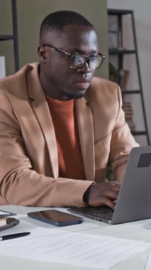 Genç Afro-Amerikan şirket müdürünün gözlüklü ve şık gündelik kıyafetleriyle masada otururken, dizüstü bilgisayarda çalışırken ve modern ofis telefonlarına cevap verirken dikey görüntüsü.