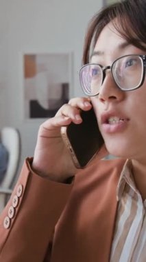 Genç Çinli kadın şirket CEO 'sunun göz giysisi ve hardal ceketi giyerken iş ortağıyla telefon görüşmesi yaparken, iş yerinde ortak başlangıç veya iş konularını tartışırken dikey yakın çekimi.