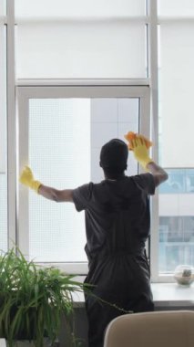 Ofis binasında çalışırken pencere camlarını kumaşla silerken, tulumlu, tanınmayan genç siyahi bir erkeğin dikey arka çekimi.