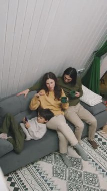 Yüksek açılı, genç anne, baba, oğul kış akşamı kanepede dinlenirken, çocuk akıllı telefonda oyun oynarken, anne bir kupa çayla saçını okşarken ve kocasıyla sohbet ederken.