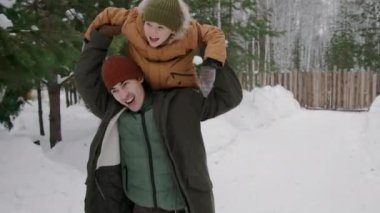 Orta boy parka montlu, yün şapkalı, eşarplı, kış tatilinde kar altında 7 yaşındaki oğluyla eğlenen, koşan, kaldıran, uçak oynayan ve gülen Asyalı bir adamın orta boy fotoğrafı.