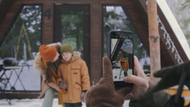 Yünlü eldivenli isimsiz bir adamın, kış tatilinde orman kulübesinin önünde sarılıp el sallayan, 7 yaşındaki bir çocukla akıllı telefonundan fotoğraf çekmesi.