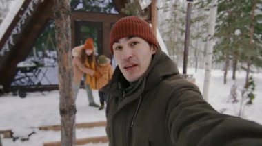 Genç Asyalı erkek blogcunun kış kıyafetleriyle sosyal medya makarasını karlı tatil kulübesinde çekerken, dondurucu oğul ve gülen eşe Noel tatili sırasında sarılırken orta ölçekli POV fotoğrafı.