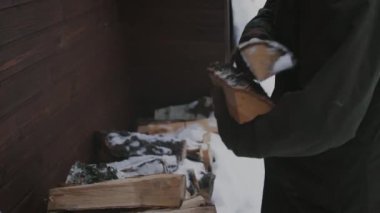 Sıcak ceketli melez bir gencin orta eğik pozu, ve örgü şapka giysileri orman kulübesinin dışındaki ahşap yığınından bir avuç kütüğü alıp şömineye ya da sobaya giderken.