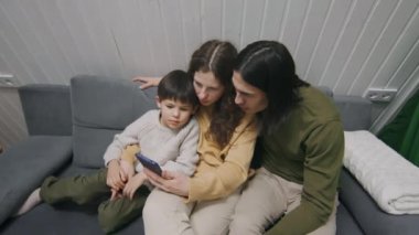 Çok ırklı bir ailenin orta-açılı görüntüsü, beyaz bir eş, Asyalı bir koca, çift ırklı bir oğul, akşamları tatil kulübesinde dinleniyor, akıllı telefondan çocuk için cep telefonu oyunu ya da çizgi film arıyor.