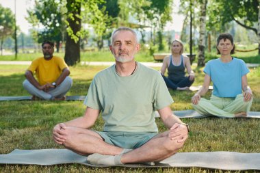 Aktif kıyafetli yaşlı bir adam, kültürler arası insanların önünde meditasyon egzersizleri yaparken bacak bacak üstüne atıyor.
