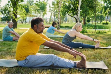 Side view of mature multiethnic man in yellow t-shirt and grey sweatpants touching toes by fingertips while exercising on mat clipart