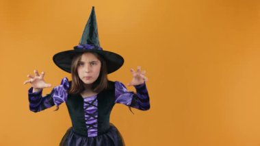 Cadılar Bayramı cadı kostümü içindeki genç büyücünün orta ölçekli görüntüsü ve kara şapkalı sihirbazlık büyüsü ve kamera karşısında boş turuncu arka planda büyücülük yapmak ve sonra da gülmek.