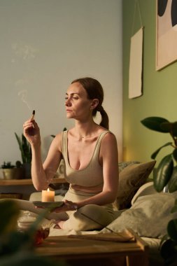 Bacak bacak üstüne atmış, yoga eğitiminden sonra koltukta dinlenirken elinde sandal ağacı çubuğu tüttüren genç, sakin bir kadın.