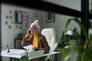 Çağdaş ev ofisinde masa başında oturan yaşlı bir kadının portresi bitkilerle ve renkli notlarla dolu, dokümanları incelerken telefon, dizüstü bilgisayar ve çalışma alanı kullanılıyor.