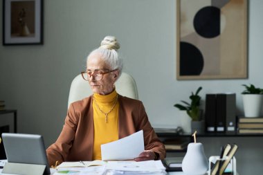 Üst düzey beyaz saçlı kadın, çalışma masasında laptop ve çeşitli ofis eşyalarıyla belgeleri incelerken moda gözlük ve bej ceket giyiyor.