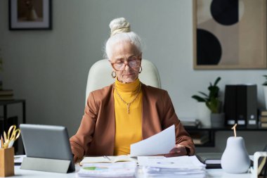 Beyaz saçlı, gözlüklü ve iş kıyafeti giyen yaşlı bir kadın masada oturuyor, raporları ve belgeleri analiz ediyor. Arka planda teknoloji ve bitkilerle birlikte modern dekor ile ofis ayarları