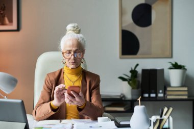 Kitaplar, belgeler ve dekorasyon eşyalarıyla çevrili bir şekilde ana ofiste otururken telefon ekranına odaklanmış gözlük takan son sınıf öğrencisi bir kadın.