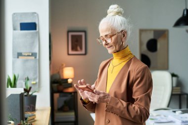 Profesyonel giysiler içinde gözlük takan, modern ofiste duran, akıllı telefonuyla meşgul olan, gülümseyen ve büyük pencerenin yanında duran son sınıf öğrencisi bir kadın.