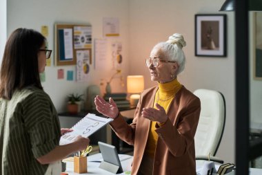 Görevde bulunan kıdemli iş kadını, elinde belgeler bulunan genç bir meslektaşla görüşüyor, takım çalışması ve profesyonel işbirliği üzerine yoğunlaşıyor, fikirler yaratıyor.