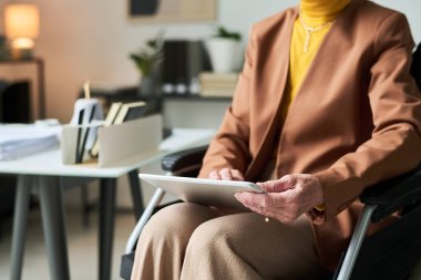 Üst düzey kadın elinde dijital tabletle ofiste oturuyor, arka planda görünen kitap ve belgelerle işine odaklanıyor ve profesyonellik duygusu katıyor.