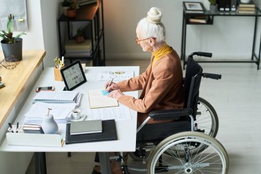 Modern bir ofis ortamında, etrafında çeşitli erzak ve bitkilerle masa başında yazan yaşlı bir kadın. Elektronik tablet kullanılarak çalıştırıldı