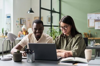 Modern bir ofis alanında dizüstü bilgisayarla uğraşan gülümseyen meslektaşlar; kitaplar, bitkiler ve sanatın yer aldığı proje üzerinde beyin fırtınası yaparken ve işbirliği yaparken