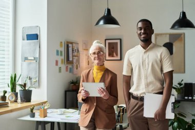 Tablet tutan yaşlı kadın ve elinde defter tutan genç adam çeşitli dekorlar ve çalışma alanlarını aydınlatan doğal ışıkla modern ofiste duruyorlar.