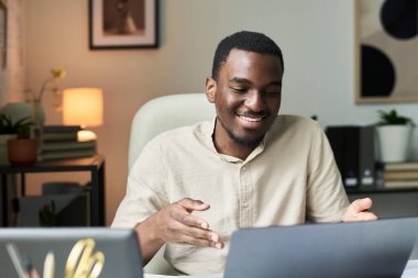 Ofiste oturan adam dizüstü bilgisayarla uğraşırken gülümsüyor. Arkaplanda modern dekor, kitaplık ve rahat bir çalışma alanı yaratan bitkiler yer alıyor