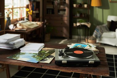 Masasında vinil oynatıcı ve istiflenmiş kitap ve evrak odasıyla birlikte retrotemalı ev ofisi. Sıcak güneş ışığı penceredeki rahat boşluğu dolduruyor ve davetkar bir ortam yaratıyor.