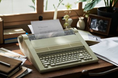 Güneş ışığı, kağıt, kitap ve bitkilerle çevrili ahşap masadaki klasik daktiloyu aydınlatıyor, rahat çalışma alanında nostaljik bir atmosfer yaratıyor.