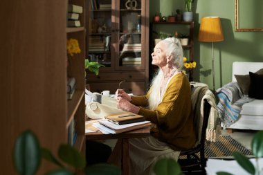 Elderly woman writing at wooden desk surrounded by bookshelves and indoor plants in well-lit, cozy room with yellow lamp and comfortable chair clipart