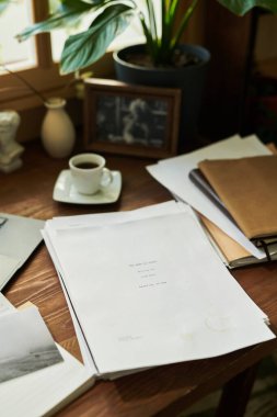 Ofis masası evrak, klasörler ve ev bitkileriyle çevrili kahve fincanı ve arka planda bulanık pencere görüntüsüyle kaplı. Çalışma alanı atmosferi yaratıyor.