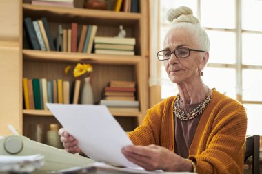 Gözlük takan yaşlı kadın portresi, ahşap masada oturuyor, elinde kağıt, arka planda kitap rafı, pencere aydınlatıcı sahnenin doğal ışığı.