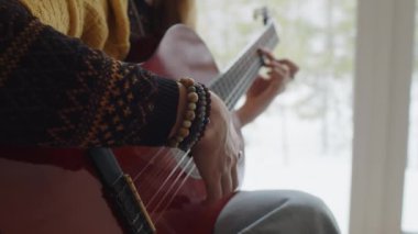 Bileğinde ahşap bileklik ve sıcak Noel süveteri olan, panoramik pencerenin yanında kırmızı akustik gitar çalan isimsiz bir müzisyenin orta bölüm çekimi.
