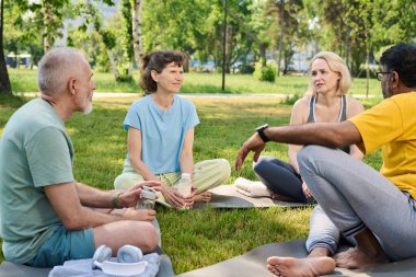 Group of intercultural mature men and women sitting on mats on green lawn in park, having water and discussing points of yoga training clipart