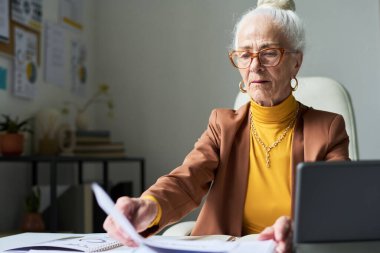 Masada oturan yaşlı kadın modern ofis ortamında belgelerle çalışıyor. Gözlük takıyor ve arka planda görünen iş çizelgeleri ve ofis malzemelerine odaklanıyor
