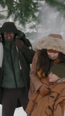 Orta boy mutlu, genç aile, Asyalı baba, beyaz anne ve melez oğulun dikey çekimi. Karların altında durup ormanın çam dallarından düşüyor ve gülüyorlar.