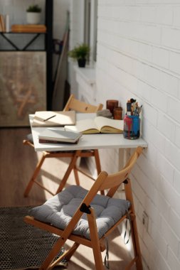 Yastıklı ahşap sandalyeler, beyaz masa ve açık defterler. Saksı bitkileri ve çeşitli yazma araçları doğal bir dokunuş ekliyor.
