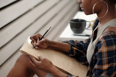 Açık havada kahve fincanı yanında otururken elinde kalemle deftere yazı yazan biri. Büyük halka küpeleri olan günlük kıyafetler giyiyordu.