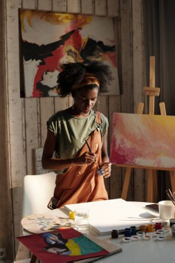 Genç ressam, soyut sanat eserleri ve çeşitli resim malzemeleriyle çevrili, parlak sanat stüdyosunda yeni bir resim üzerinde çalışırken, doğal ışık odayı dolduruyor.