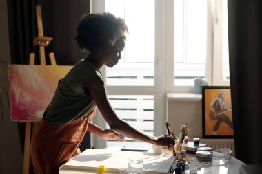 İyi ışıklandırılmış bir stüdyoda çalışan genç bir sanatçı. Etrafı pencerelerle çevrili. Doğal ışık sağlıyor ve huzurlu bir atmosfer yaratıyor.