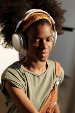 Afrika kökenli Amerikalı kadın kulaklık takarken gülümsüyor, modern kapalı mekan ortamında müziğin tadını çıkarıyor. Güneş ışınları uzayı aydınlatırken saçlar saç bandıyla bağlanmış.