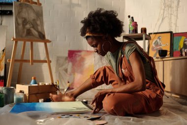 Afro-Amerikalı kadın, fırçalar, tuvaller ve yaratıcı malzemelerle çevrili rahat bir stüdyonun zemininde otururken resme dalmıştı.