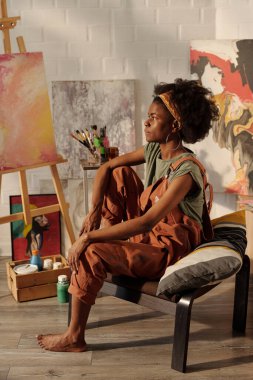 Genç Afrikalı Amerikalı kadın sanatçı, sanatsal malzemelerle sandalyede oturuyor, parlak, samimi stüdyo ortamında soyut resimler yapmaya odaklanıyor.
