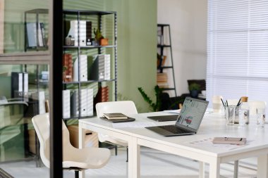 Dizüstü bilgisayarı, defterleri ve raflarında saksı bitkileri olan modern bir ofis. Perdelerin arasından süzülen güneş ışığı çalışma alanını aydınlatıyor