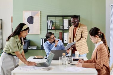Ofis ekibi, takım üyesi Standing 'in tuttuğu tablete odaklanarak iş stratejisini tartışıyor. Çeşitli takımlar tartışmalara girerken kağıtlar ve laptoplar masada.