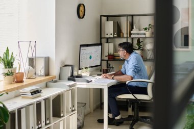 Üst düzey adam modern ofis ortamında bilgisayar verilerini analiz ediyor raflar, bitkiler ve dekorasyon ürünleri. Yüzyılın ortalarında modern stil odada ahenkli bir şekilde hissediliyordu.