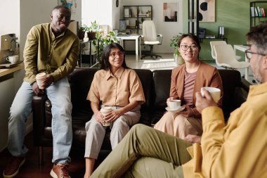 Bir grup farklı iş arkadaşı bir kanepede oturuyor ve modern ofiste rahat bir atmosferle fikir alışverişinde bulunuyor, kahve fincanları tutuyor ve sohbet ediyorlar.