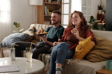 Fast food 'u olan genç ve neşeli bir çift oturma odasında rahat bir kanepede dinleniyor ve meraklı TV yayını ya da film izliyor.