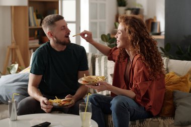 Mutlu genç bir kadın, erkek arkadaşının ağzına patates kızartması koyarken hem evinde kanepede oturuyor hem de fast food yiyor.