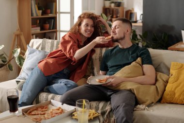Oturma odasındaki kanepede yemek yerken erkek arkadaşının ağzına pizza koyan mutlu kız.