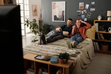 Bir kase patlamış mısırı olan genç adam televizyon kanallarında film listesini kaydırırken uzaktan kumandada düğmelere basıyor.