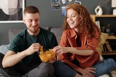 Keyifli kız ve erkek, boş vakitlerinde yatakta oturup televizyon ya da film izlerken kaseden patates cipsi alıyorlar.
